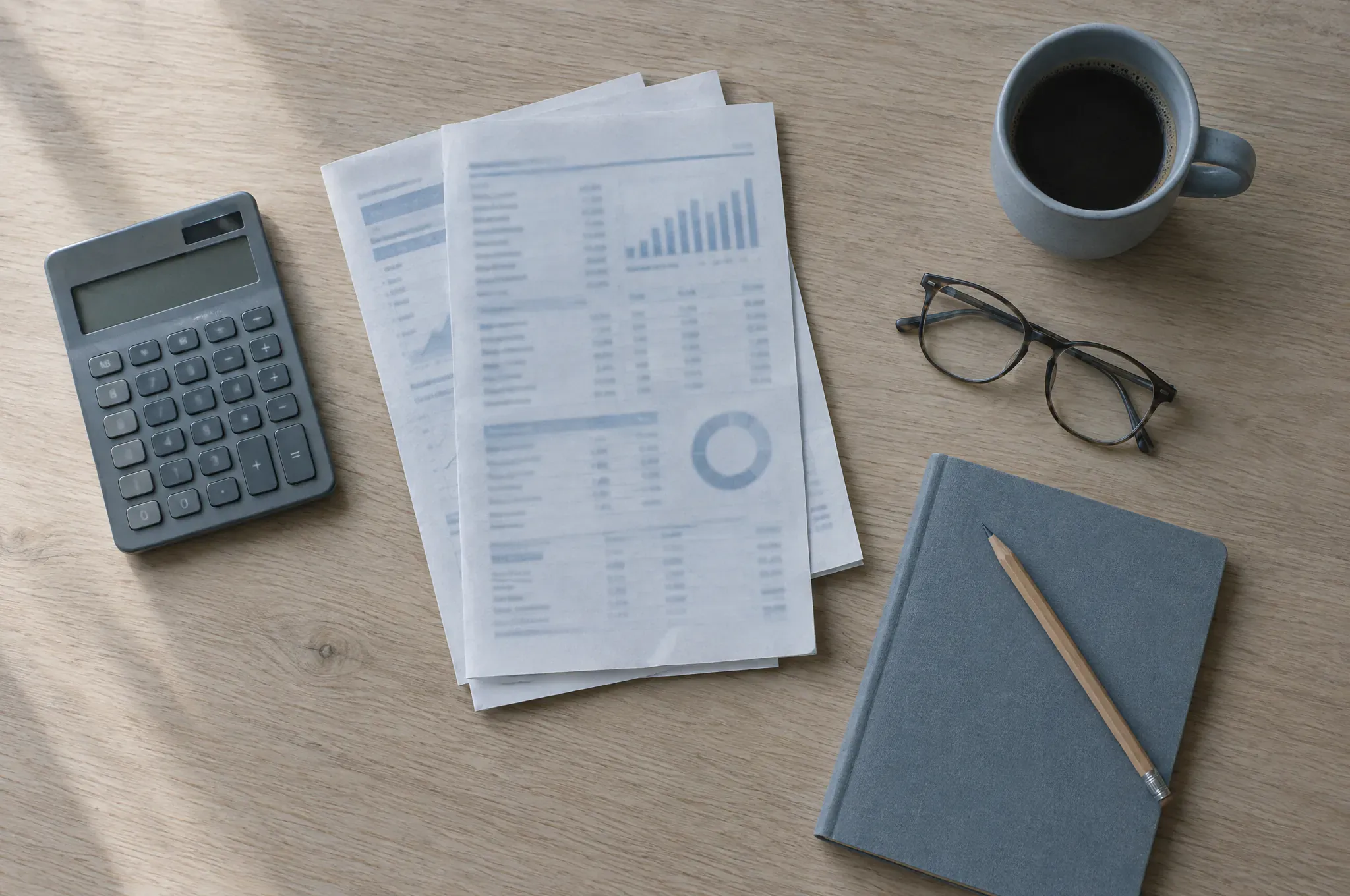 A calculator, printed financial statements, reading glasses and a notebook on an oak table.