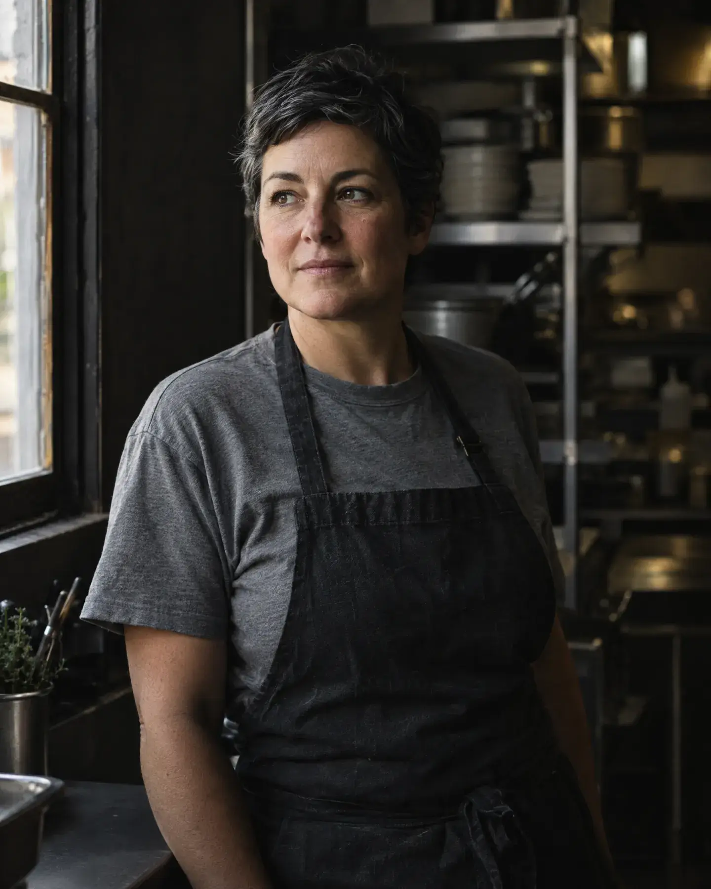 Chef Nora Kettleby in a dark apron and grey T-shirt stands by the kitchen window, looking out.