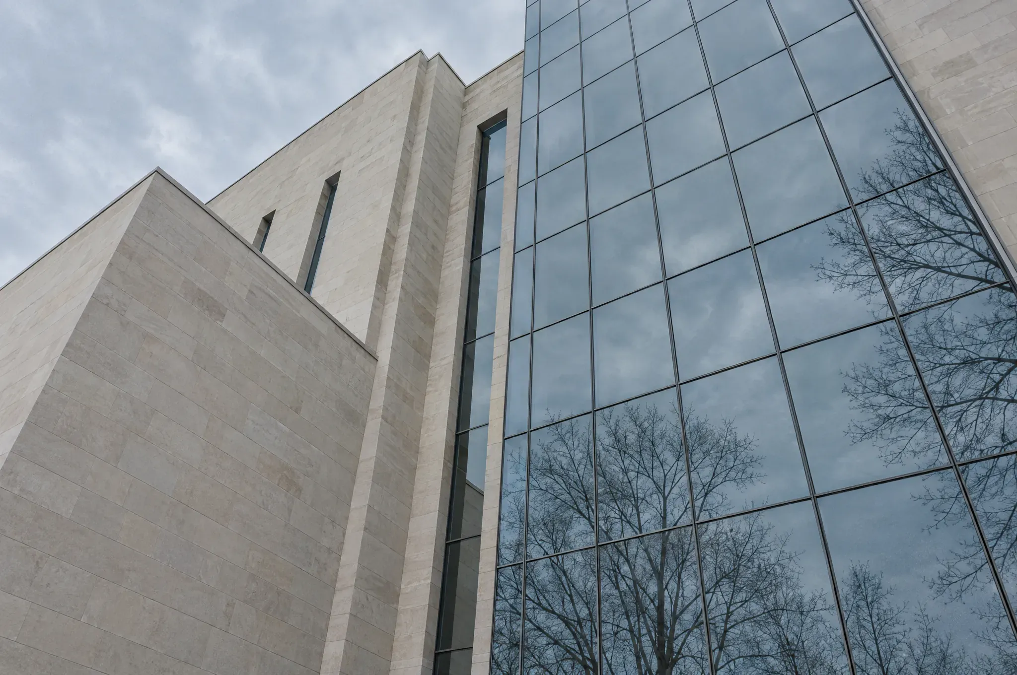A stone and glass civic building seen from below, reflecting an overcast sky and bare branches.