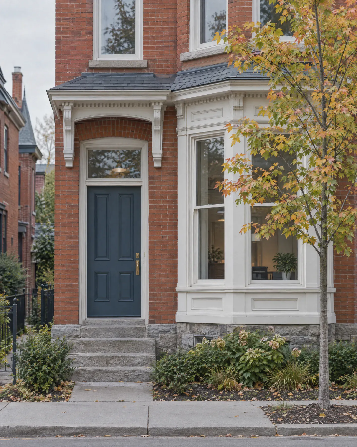 The slate blue front door of a restored red brick Victorian house, with white trim and a young maple beside the steps.