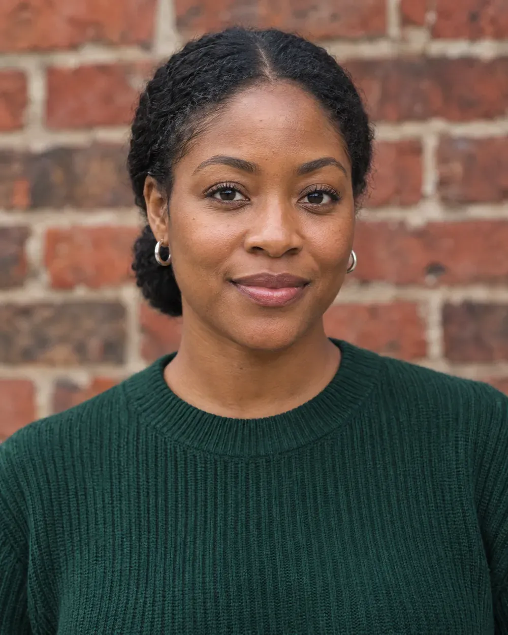 Keziah Moreau, a woman with her hair in a low bun, in a green ribbed sweater, against a brick wall.