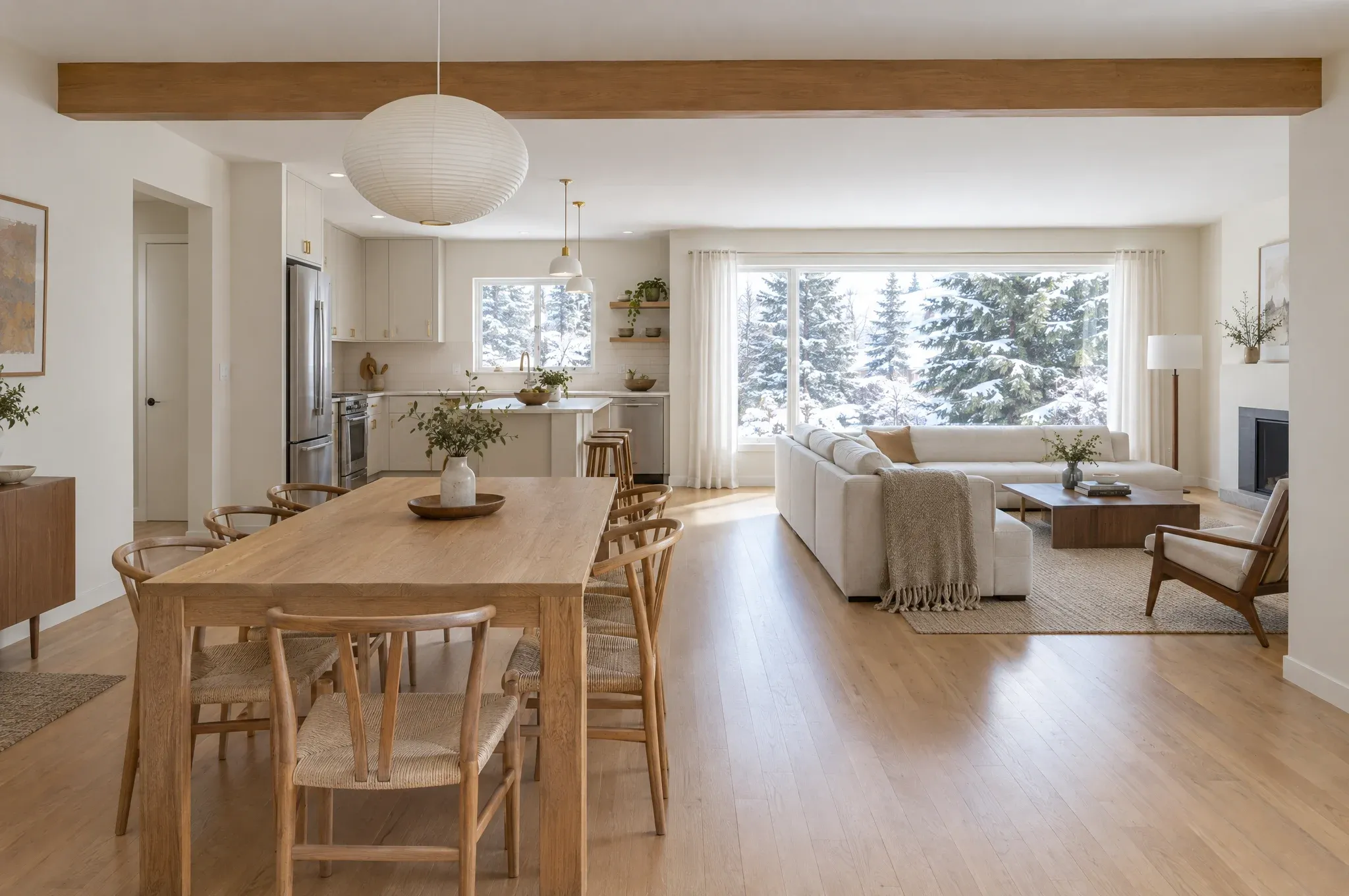 An open main floor in a bungalow, with an oak dining table, a light kitchen and a living area facing snowy evergreens.