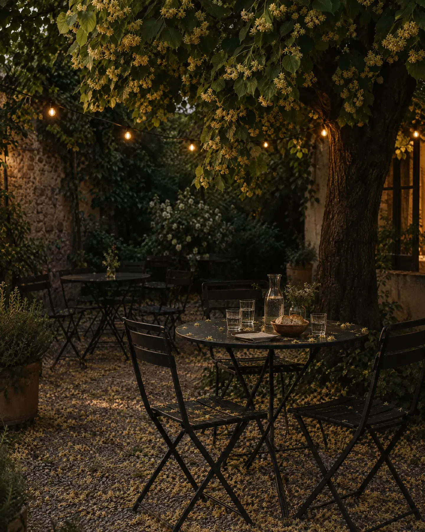 The patio under a blooming linden at dusk, with black metal bistro tables, string lights and blossom on the gravel.