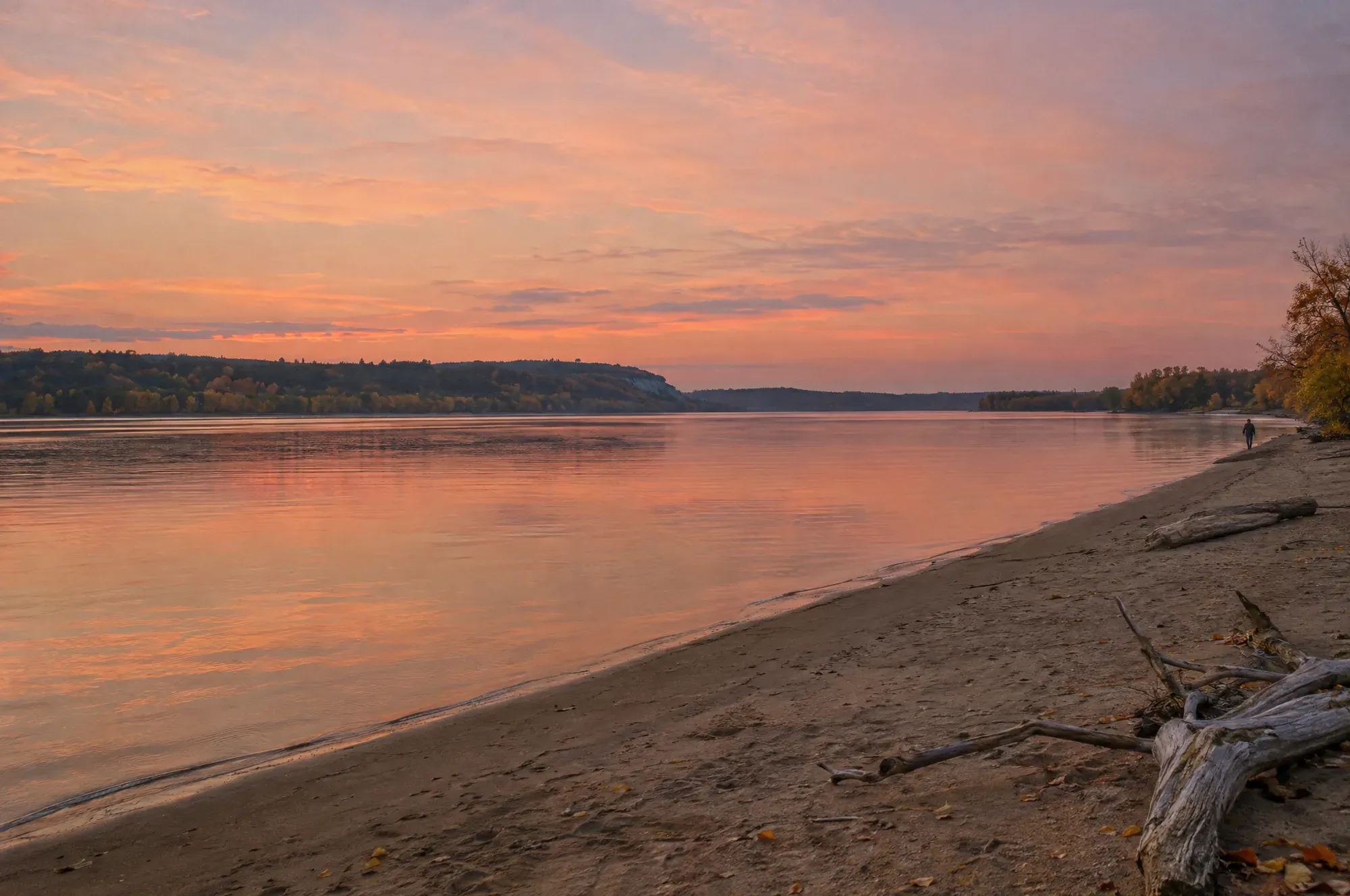 A calm river at dusk under a rose sky, with one person walking along a sandy shore in the distance.