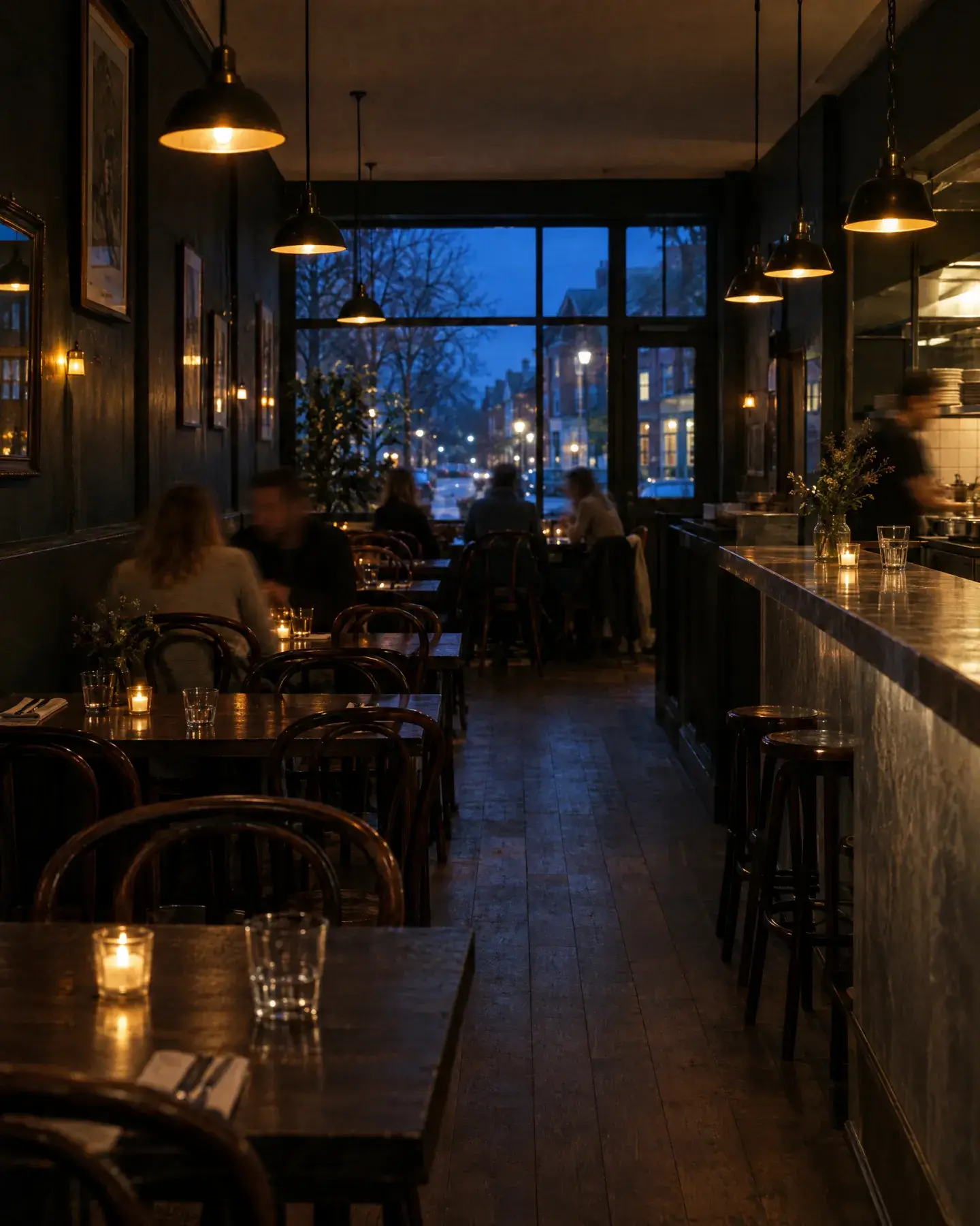 The narrow dining room at dusk, with bentwood chairs, candles on dark tables, pendant lamps over a zinc counter and diners by the front window.