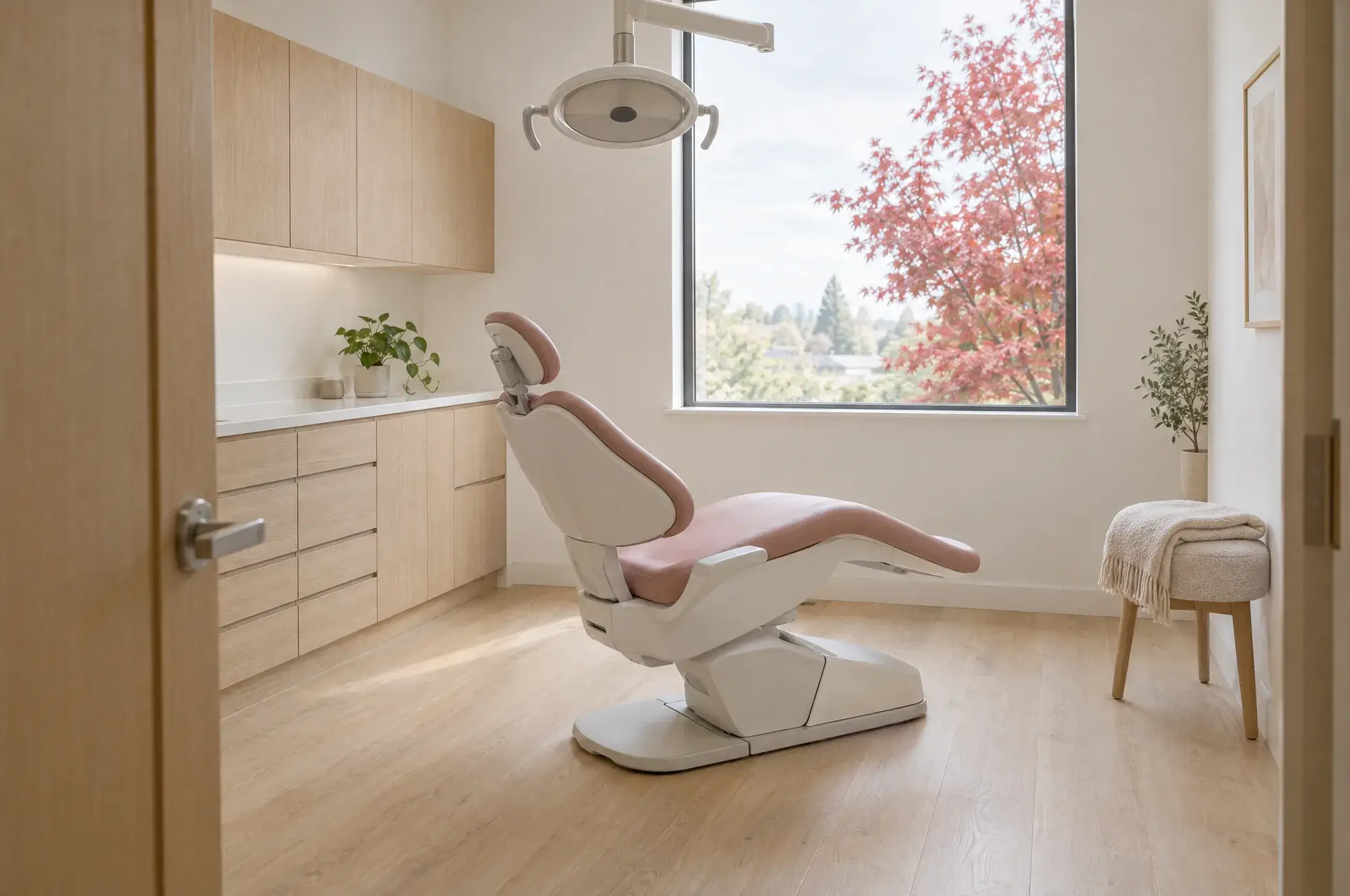 An empty treatment room seen from the doorway. A white chair with a pink seat, oak cabinets, a folded blanket on a stool and a tall window onto a red maple.