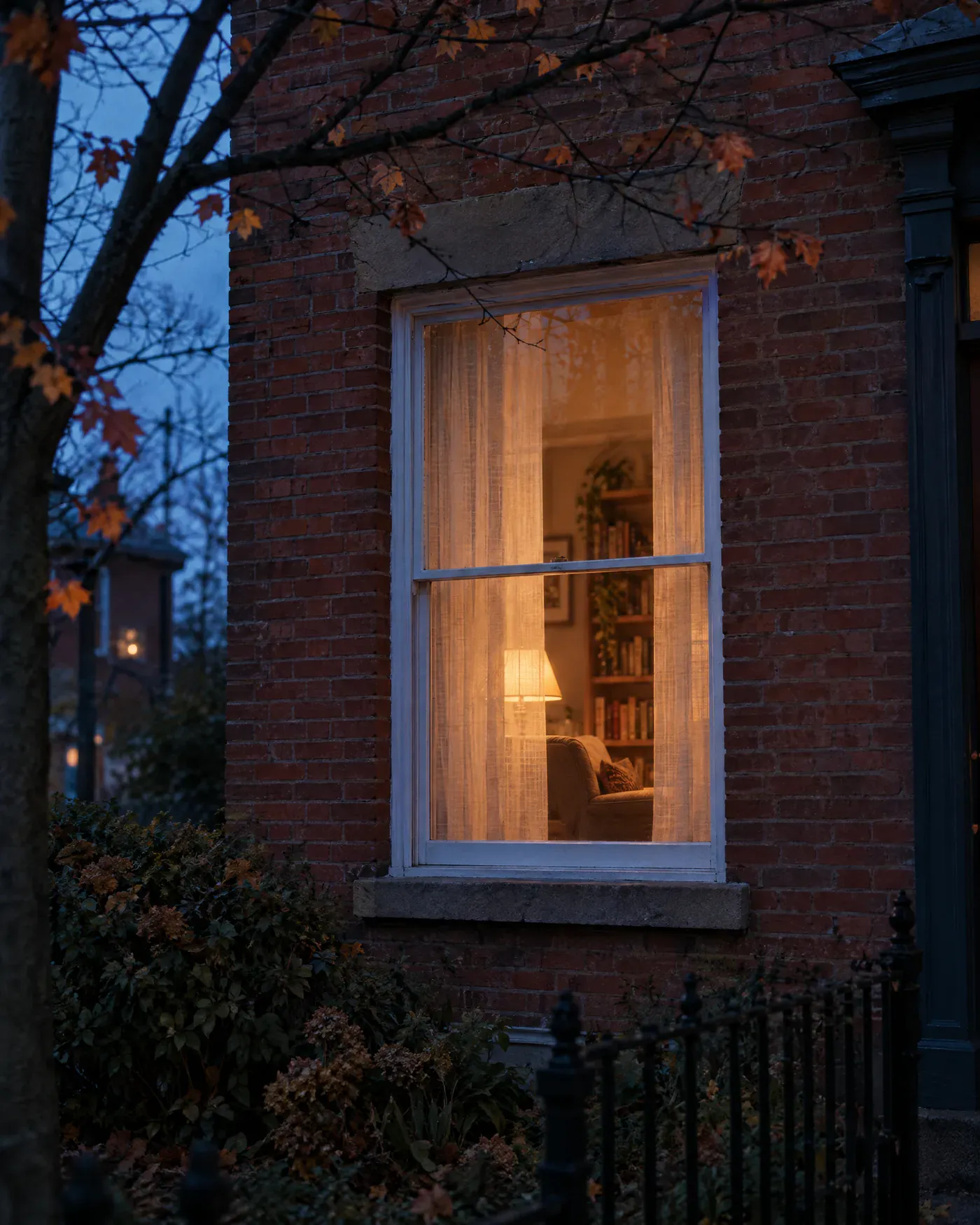 A lit window in a red brick house at dusk, with an armchair and a bookshelf glowing behind a linen curtain.